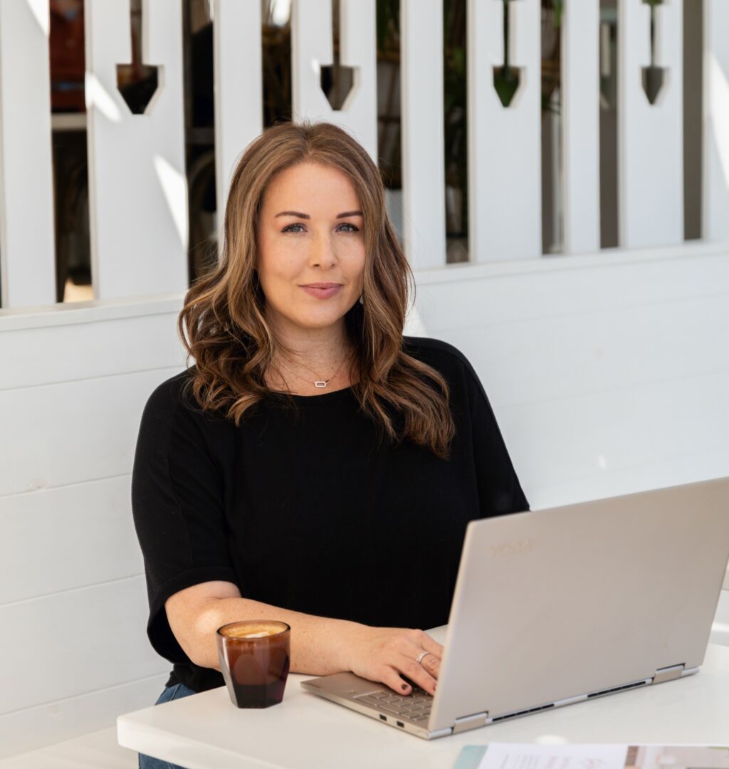 Woman with wavy hair sitting at a table with a laptop and a glass, in front of a white fence.