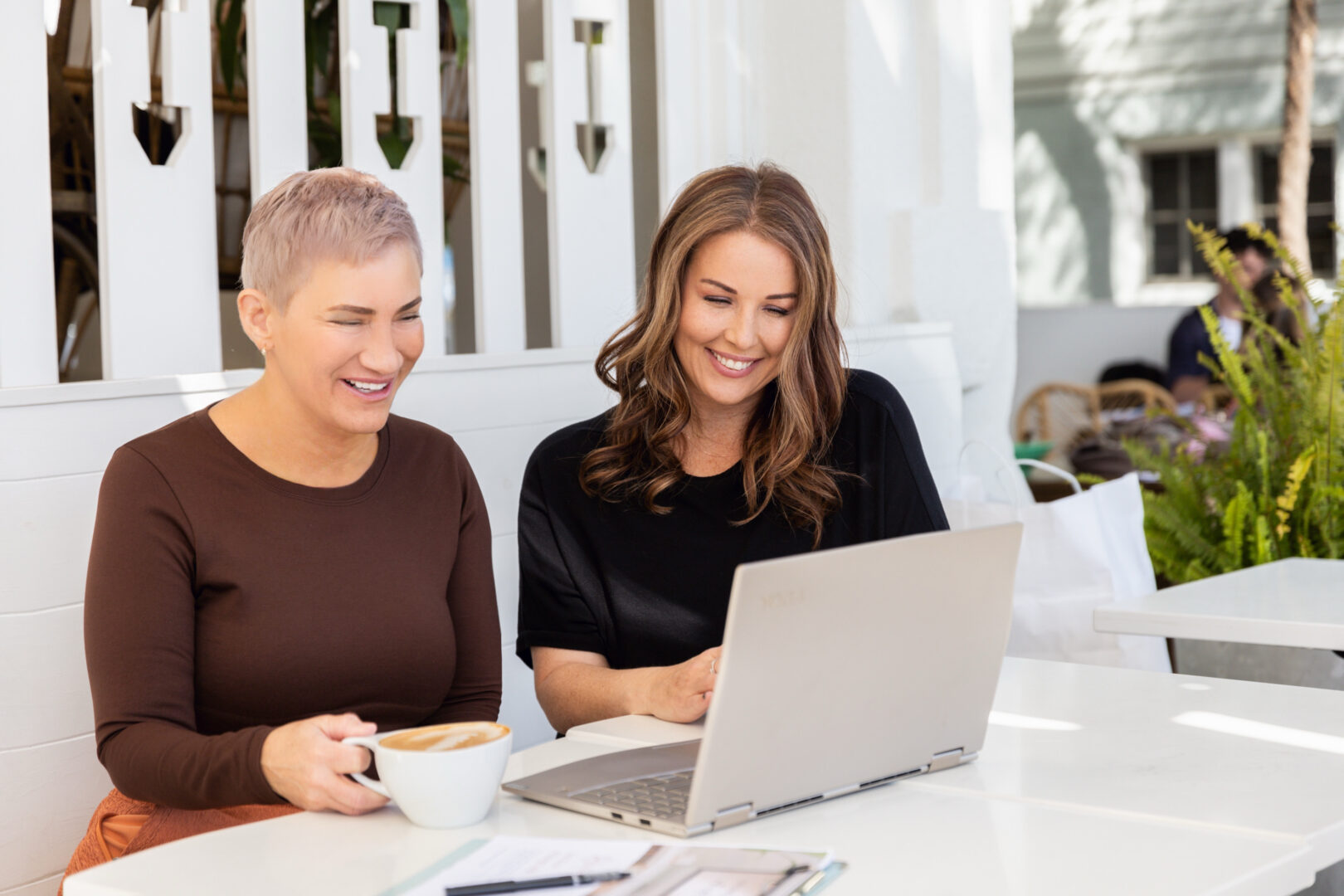 Two women sitting at a white table outdoors, smiling at a laptop, one holding a bowl.