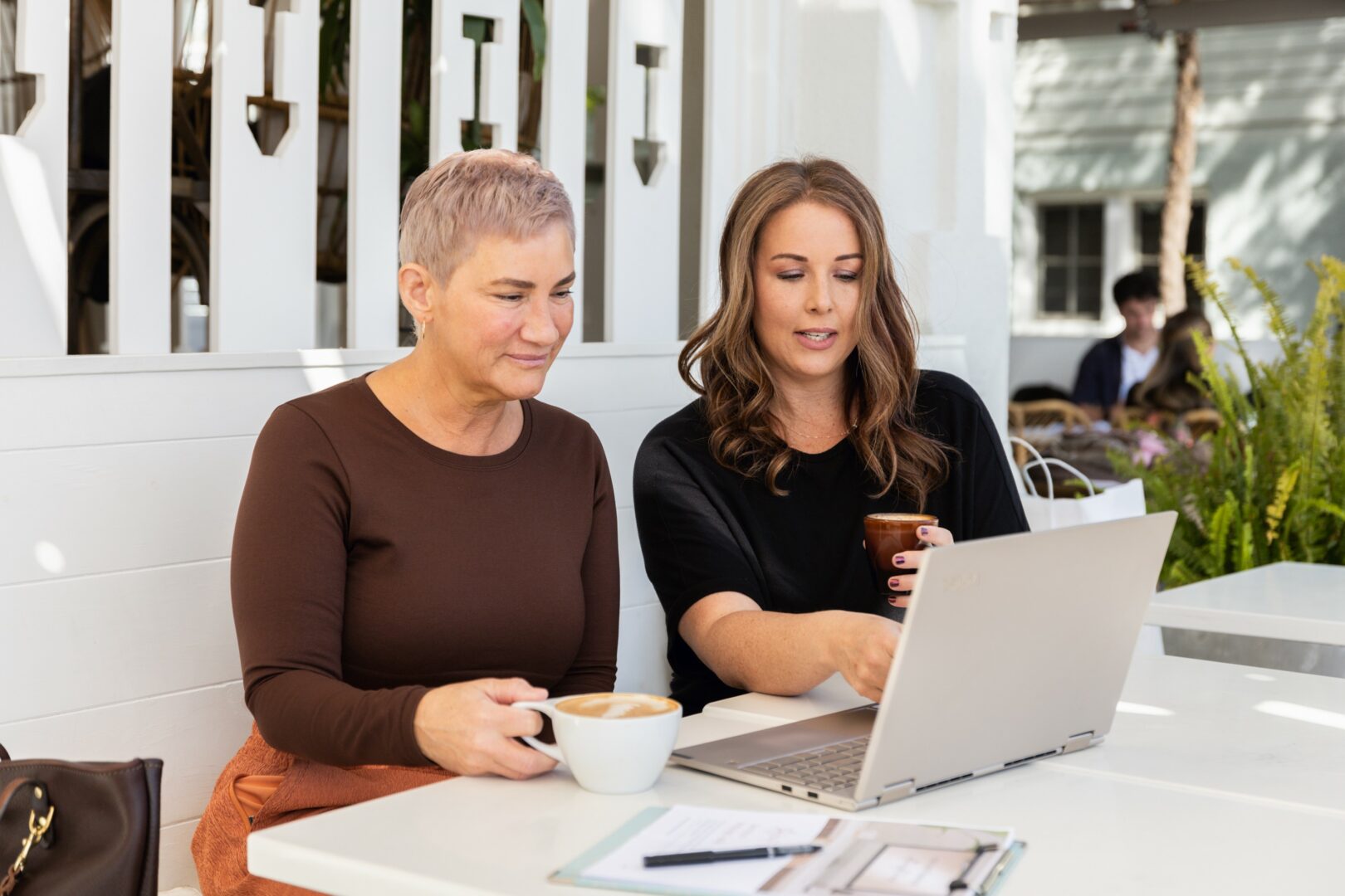 Two women sit at a white table outdoors, one with a laptop and the other holding a cup, engaged in conversation.