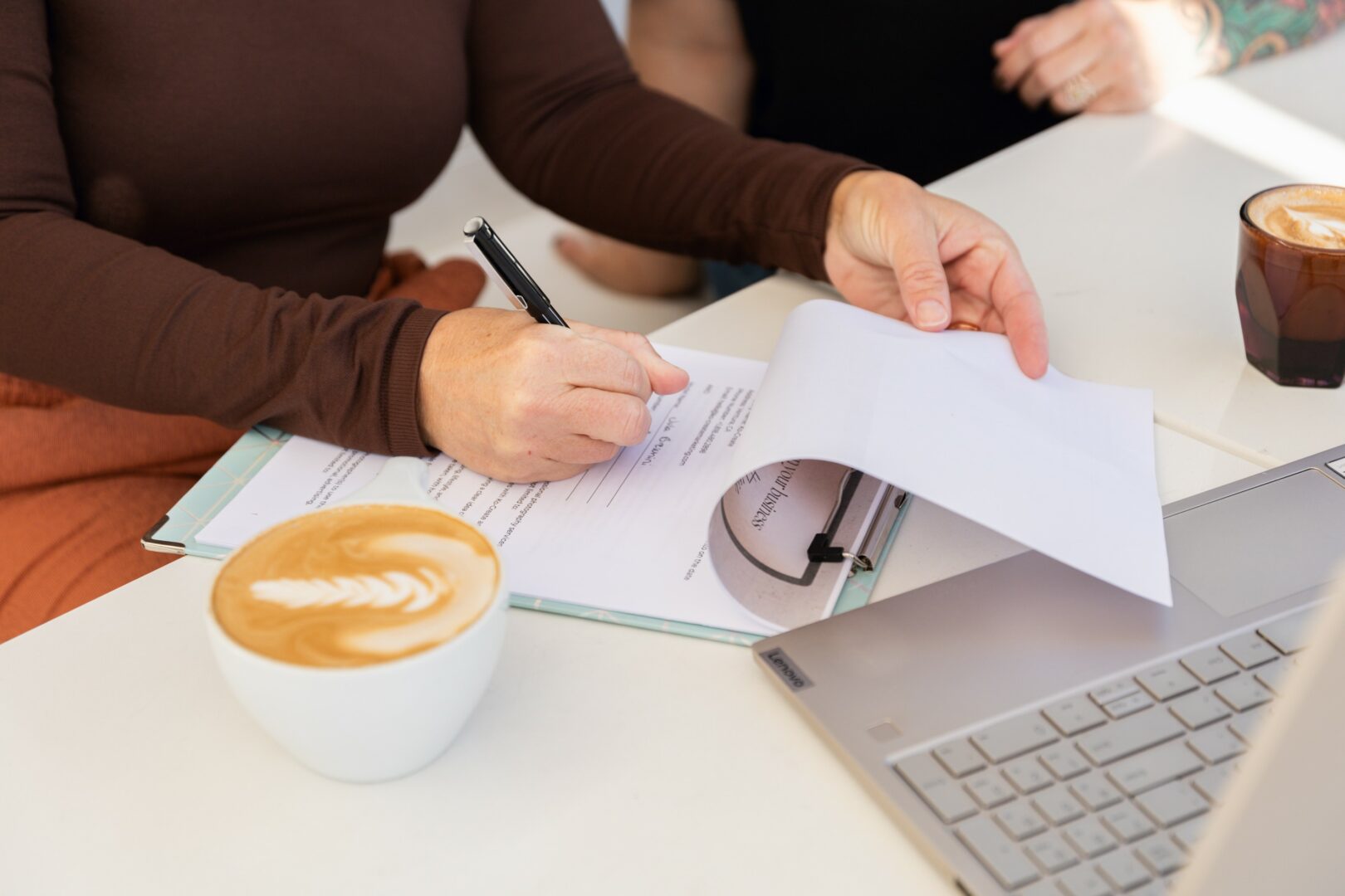 Person writing on paper with a pen, two cups of coffee, and a laptop on a white table.