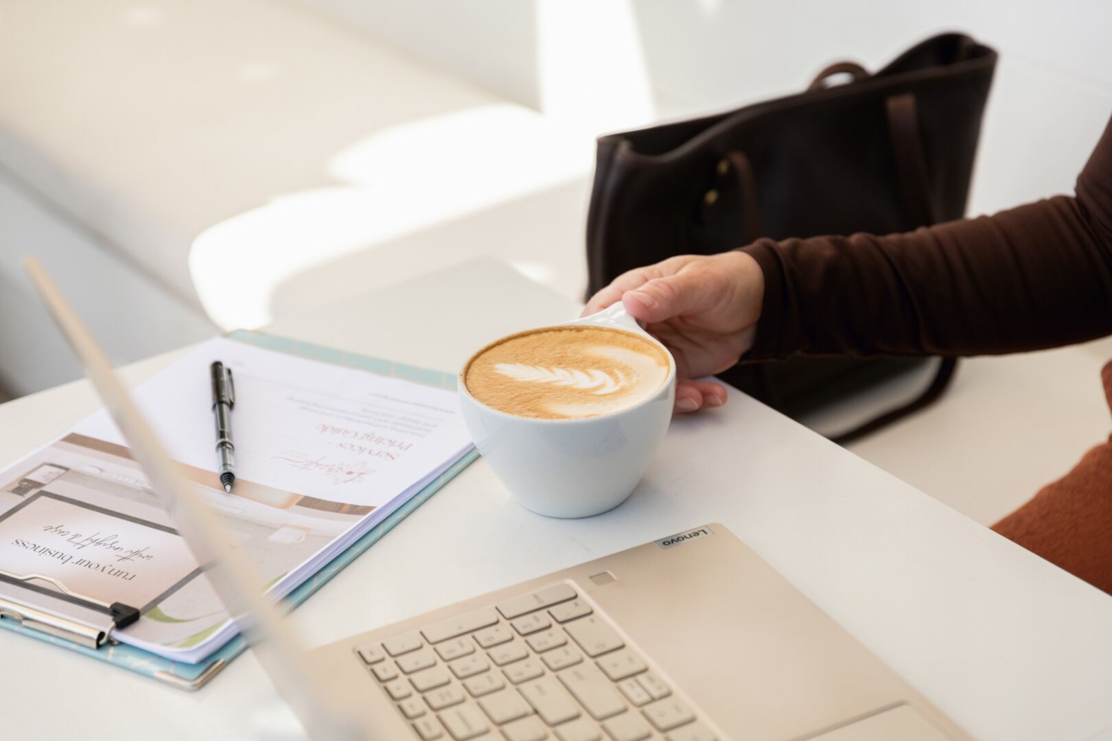 Person's hand reaching for a cup of coffee on a white table with a laptop, notebook, and pen.