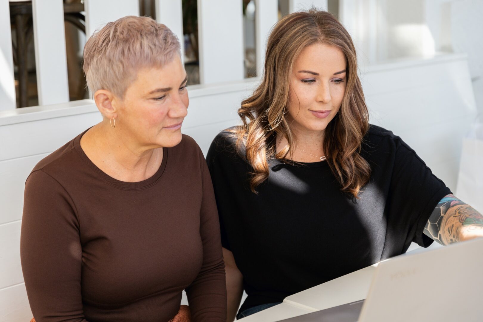 Two women sitting side by side looking at a laptop screen, one with short gray hair and the other with long brown hair.