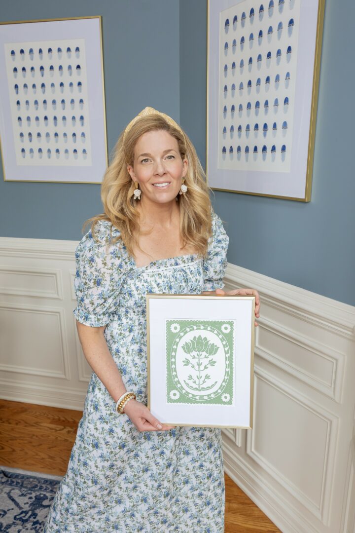 Woman with blonde hair in floral dress holding framed artwork, standing in art gallery with blue walls and framed prints.