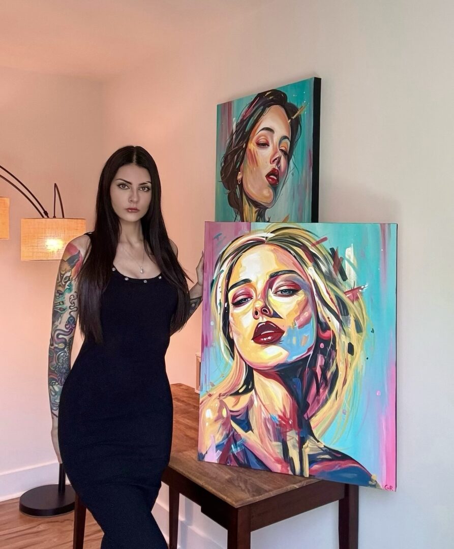 Young woman with long dark hair and tattoos on her arms standing indoors next to colorful portrait paintings on a table.