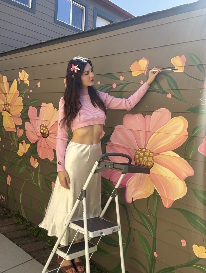 Young woman with long dark hair, wearing pink crop top and cream skirt, painting flowers on a wall mural.