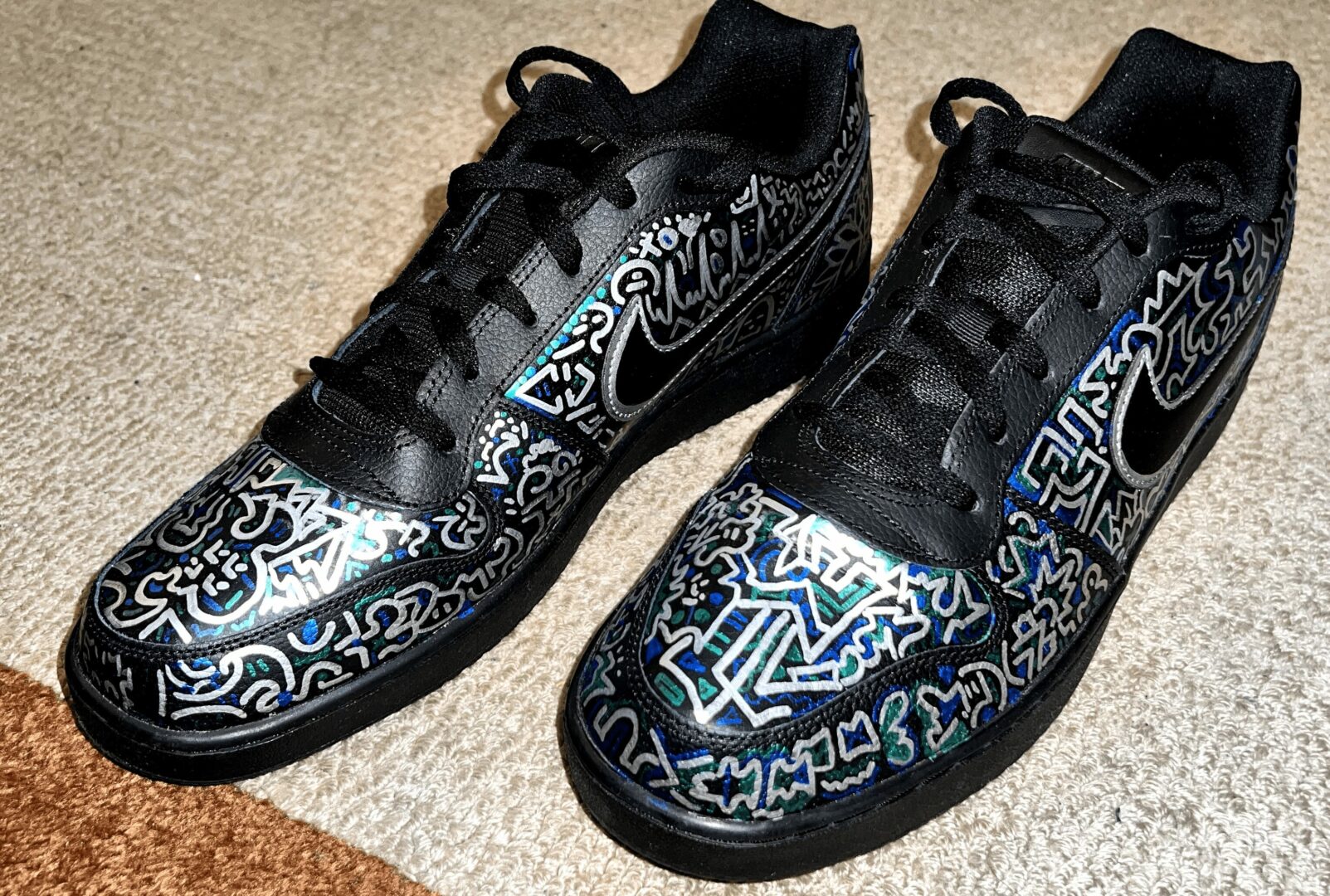Pair of black sneakers with colorful abstract patterns on the toe caps and sides, laced and placed on carpet.