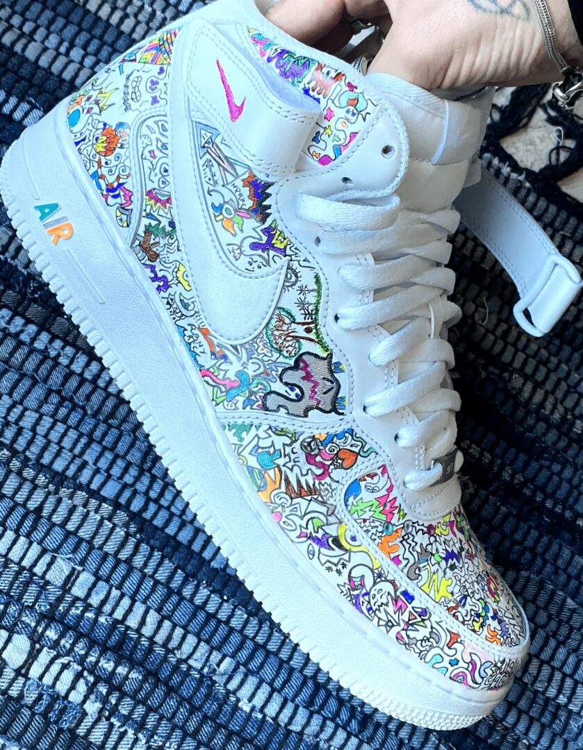 White sneaker with colorful, intricate doodle designs and a pink Nike swoosh logo, on a textured blue surface.