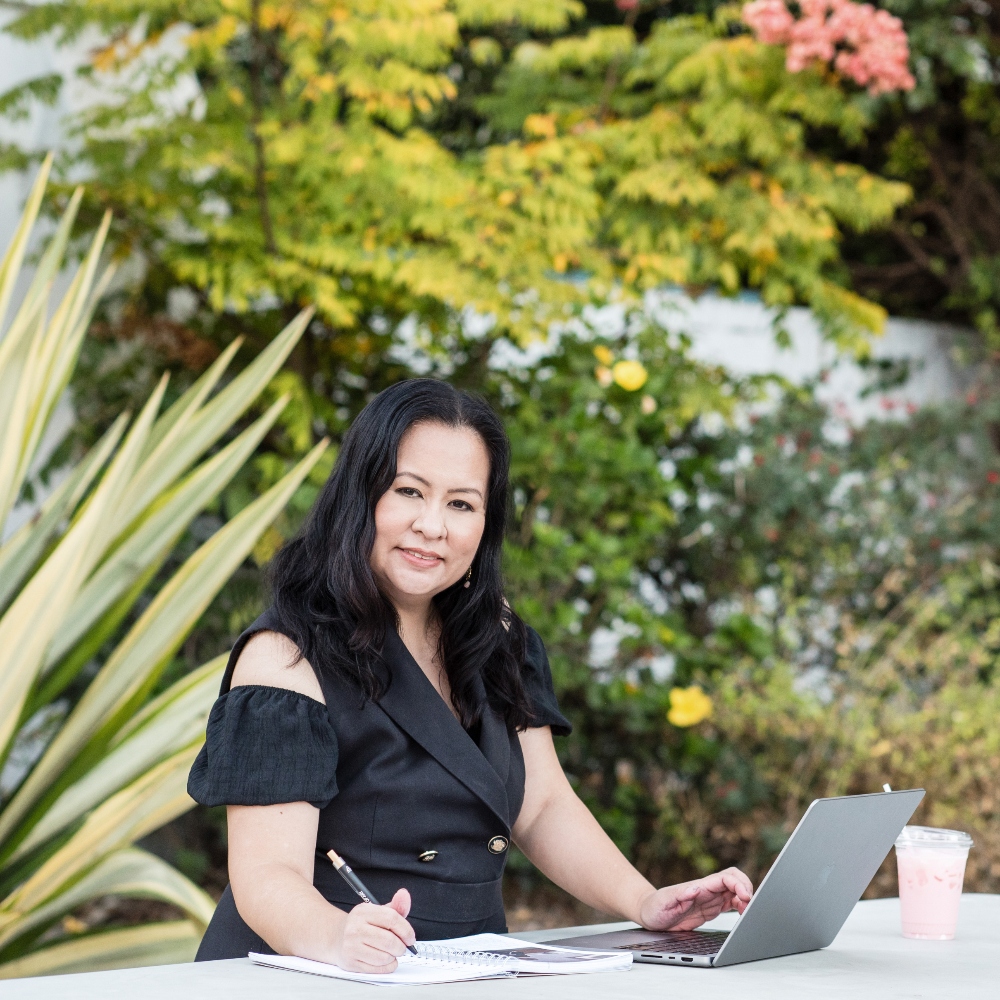 Woman sitting outdoors with a laptop and notebook, surrounded by green and yellow foliage.