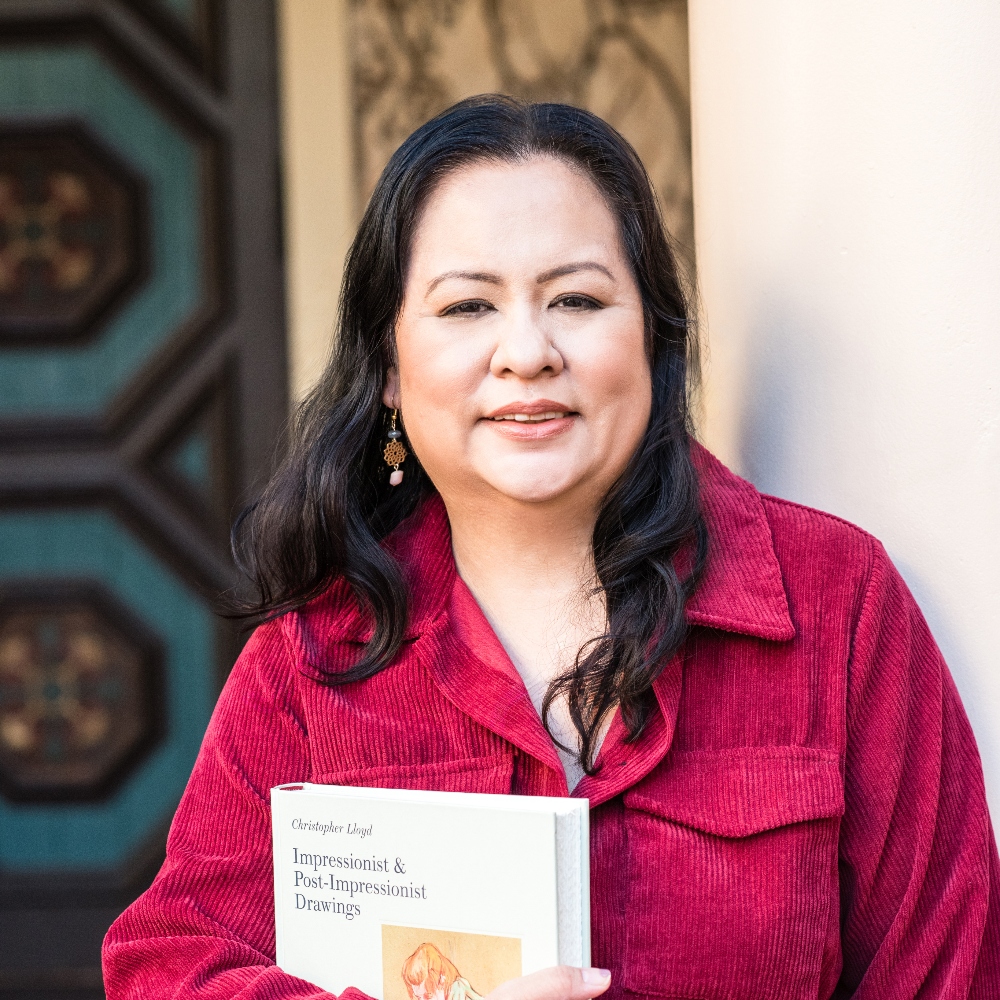 Woman with dark hair wearing a red jacket, smiling outdoors, holding a book.