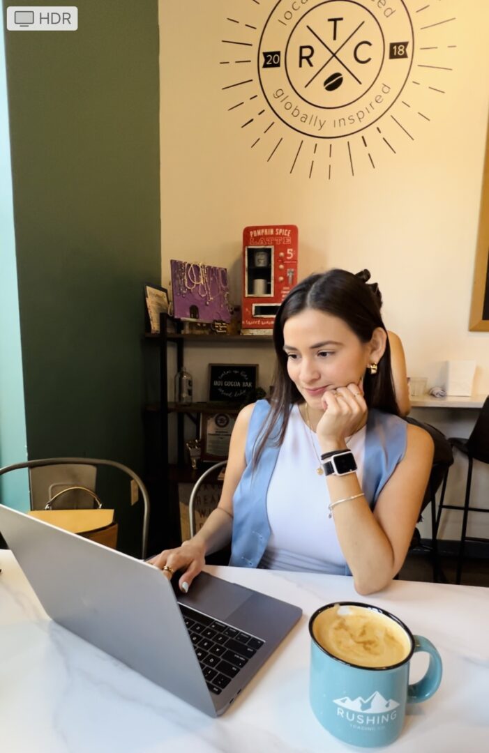 Young woman with long dark hair working on a laptop at a table in a cafe, with a blue mug nearby.
