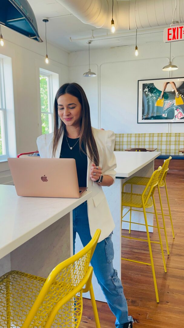 Woman working on a laptop at a white counter in a bright, modern cafe with yellow chairs and artwork on the wall.