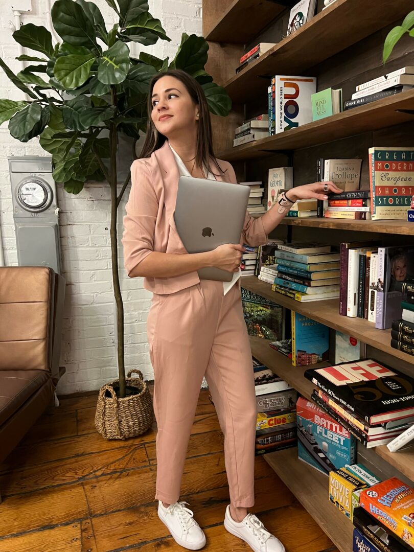 Young woman in pink jumpsuit takes a selfie with a laptop in a library, surrounded by bookshelves and a potted plant.