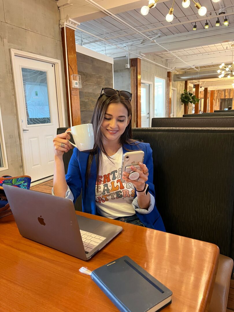 Woman sitting at a table with a laptop, holding a glass, in a cozy indoor setting.