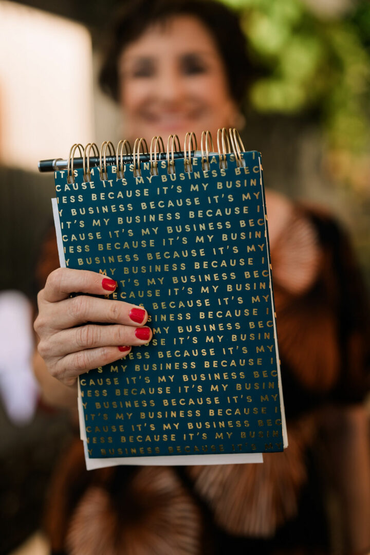 Person holding a blue notebook with white text, blurred face, outdoors background with greenery.