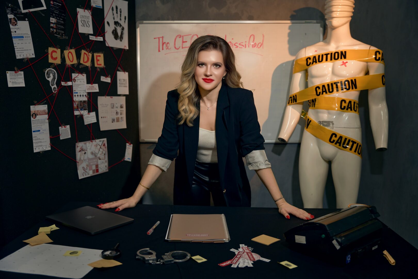 Woman with wavy hair in a blazer stands behind a desk with papers and laptops, in a room with a mannequin wrapped in caution tape.