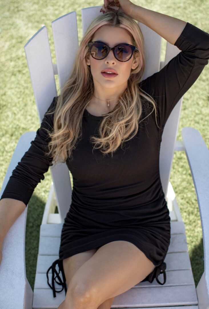 Woman with long hair wearing sunglasses and black dress sitting on a white outdoor chair.