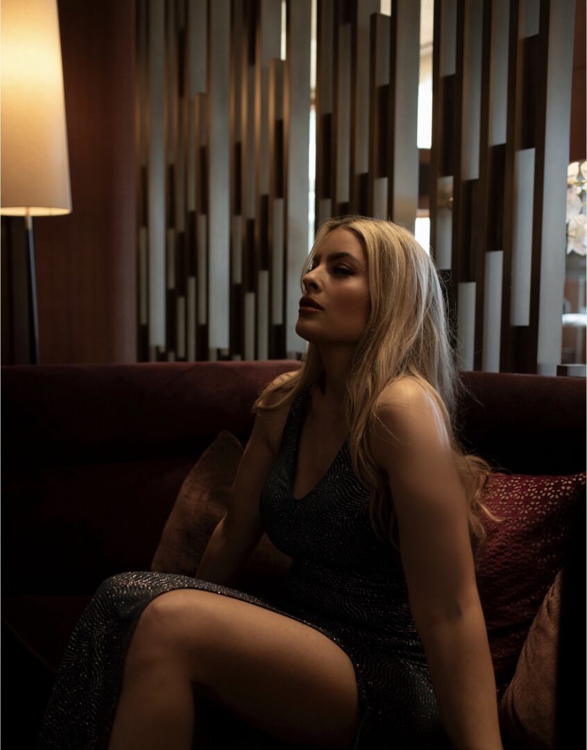 Woman with long blonde hair sitting on a sofa in a dimly lit room with patterned curtains.