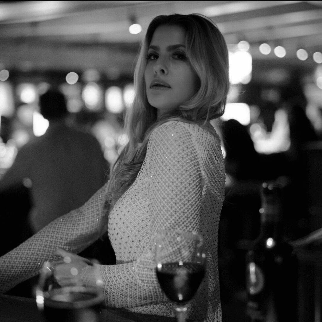 Woman with long hair in a patterned top, sitting in a dimly lit setting with wine glass nearby.