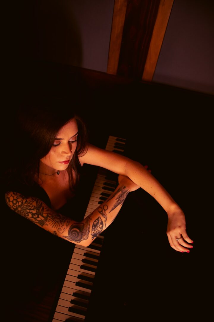 Woman with tattoos playing piano in dimly lit room, looking down at keys.