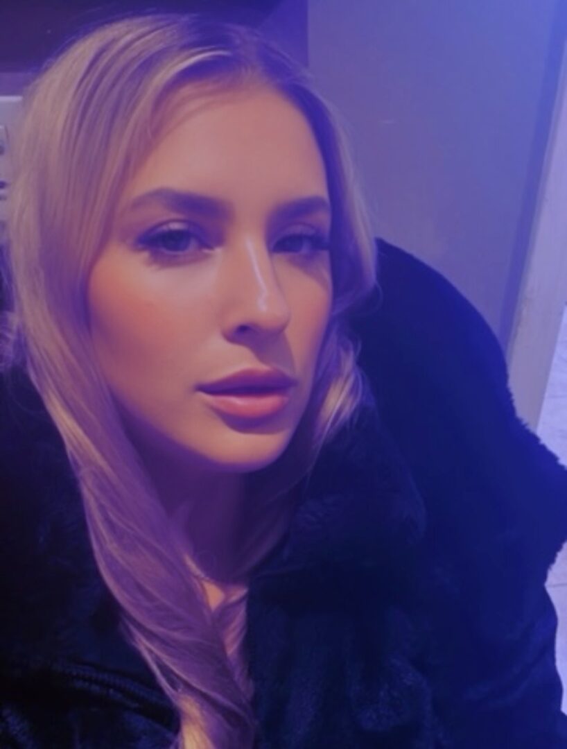 Close-up of a woman with long blonde hair, looking at the camera, with purple lighting in the background.