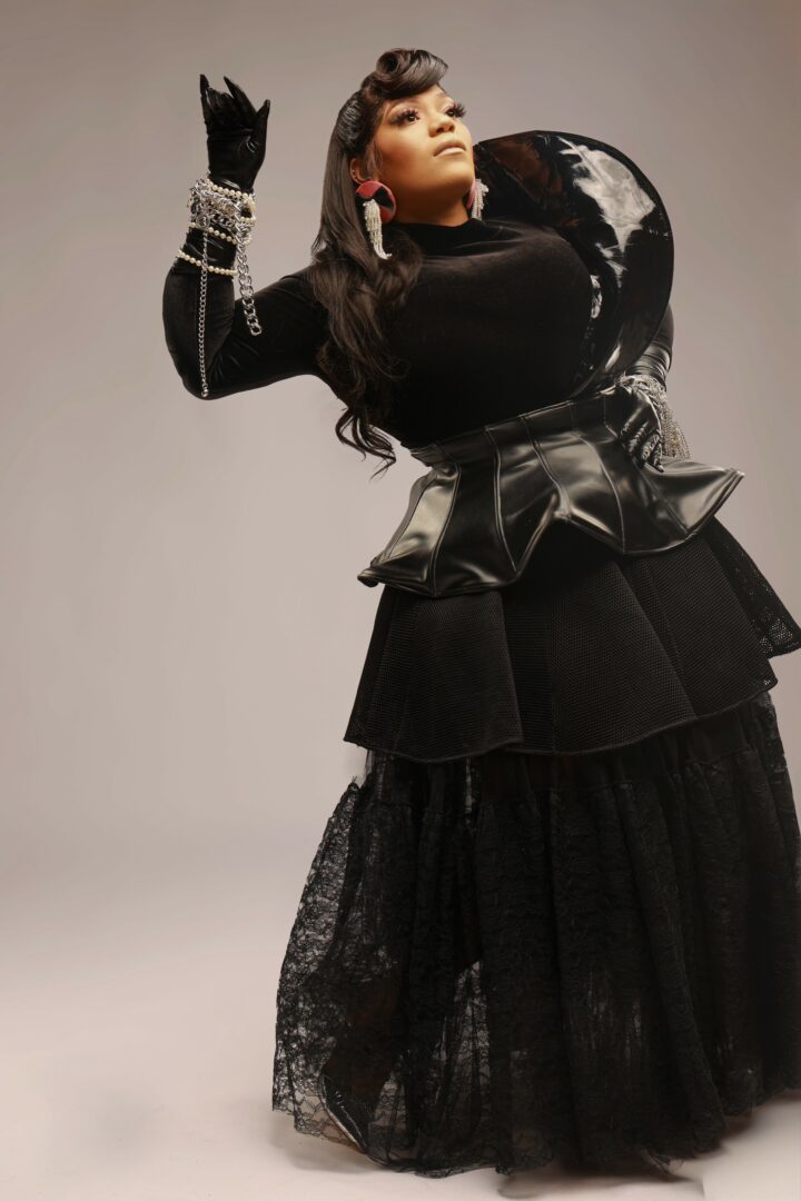 Woman in black outfit with jewelry and earrings, holding a large black bag, posing against a plain background.