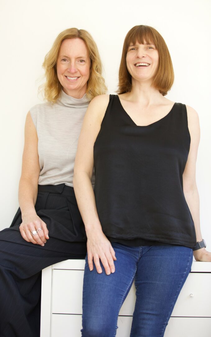 Two women smiling, standing close, one in a sleeveless black top and jeans, the other in a sleeveless gray top and dark pants.