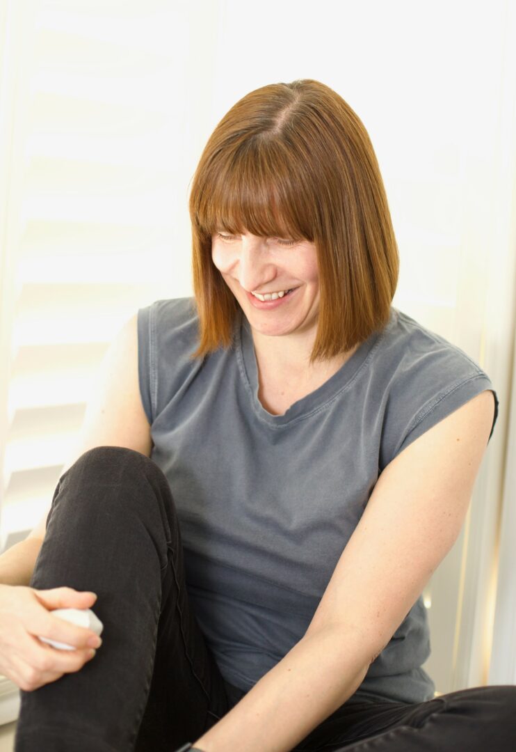 Woman with shoulder-length brown hair smiling, wearing a gray sleeveless top, sitting indoors.