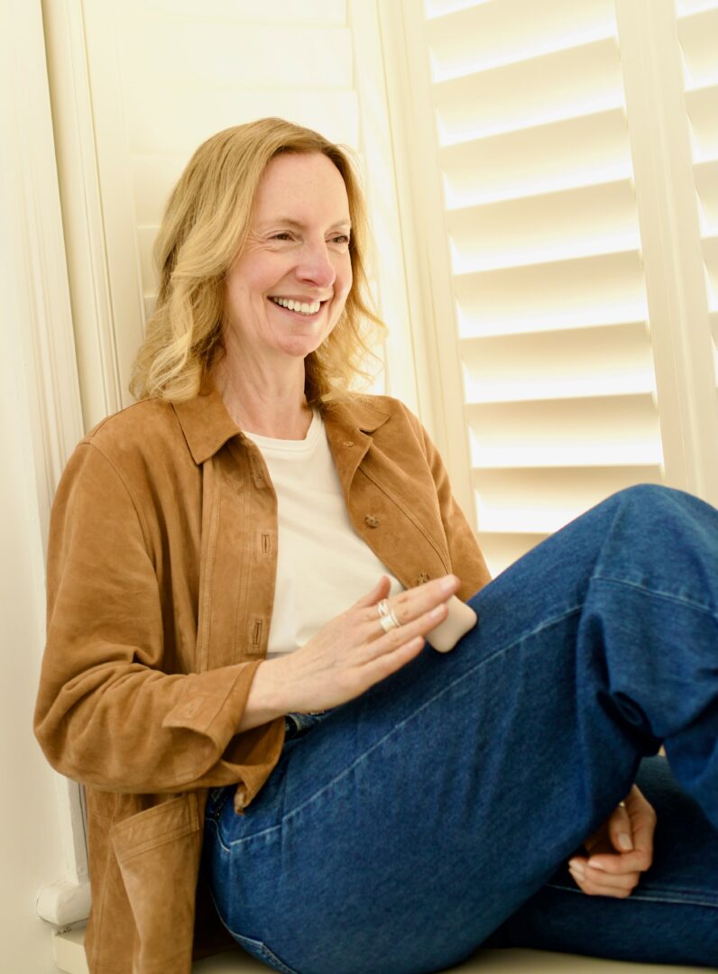 Woman with blonde hair smiling, sitting near window with white shutters, wearing a brown jacket and white shirt.