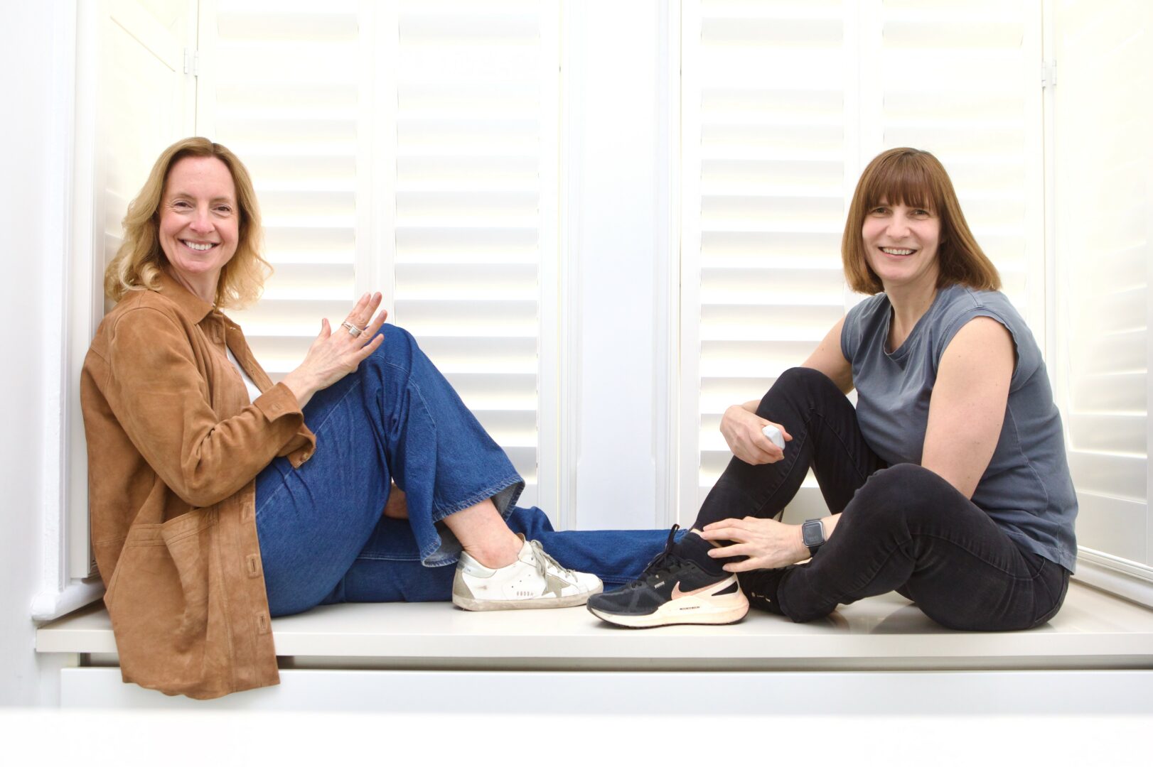 Two women sitting on a windowsill, smiling, with one resting her foot on the other's knee, in front of white blinds.