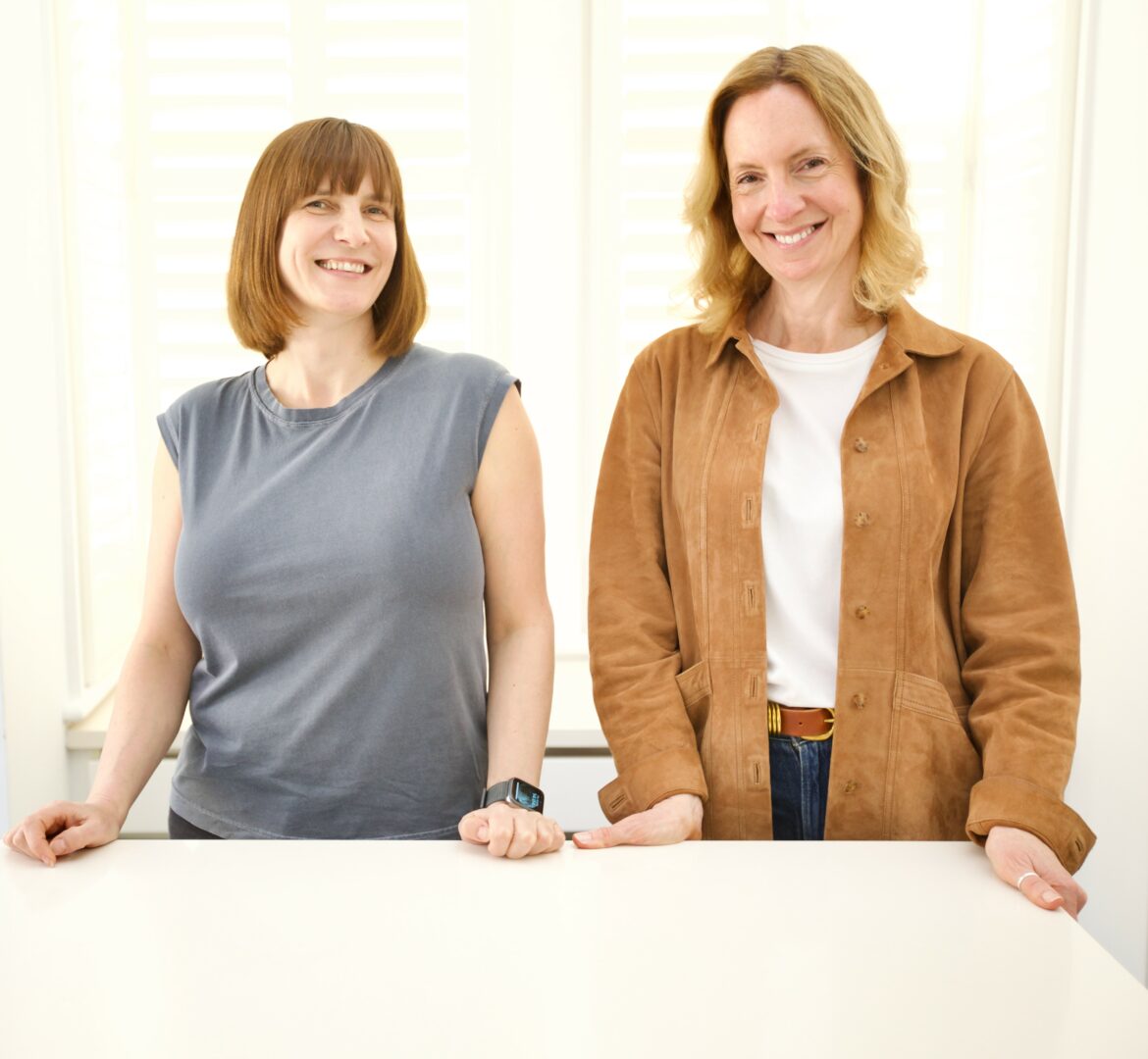 Two women standing behind a white table, smiling, in a bright room with white blinds.