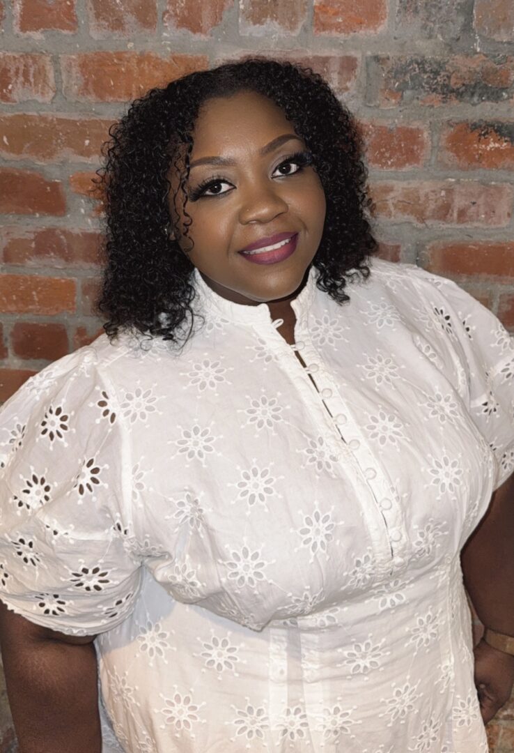 Woman with curly hair wearing a white embroidered blouse, standing against a brick wall.