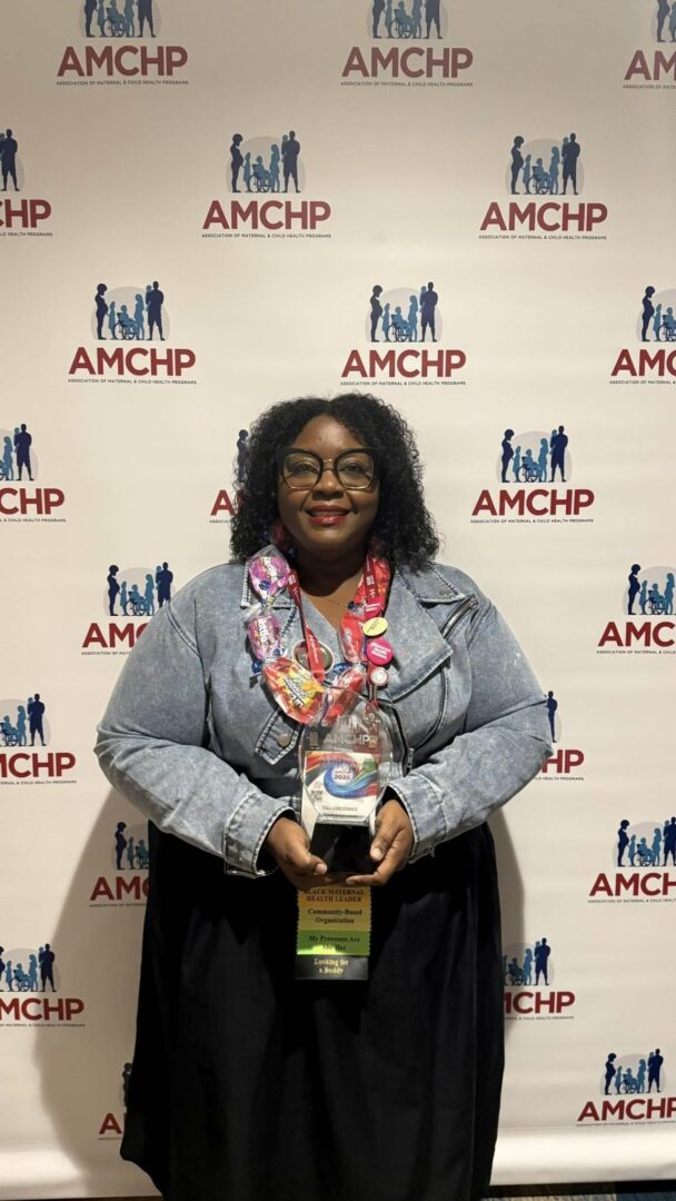 Woman with curly hair and glasses holding an award, standing in front of a backdrop with AMCHP logos.