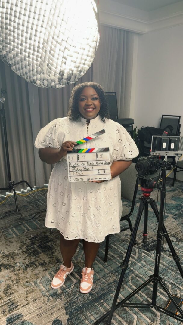 Woman in white dress holding a clapperboard, smiling, standing in a room with camera equipment and a large hanging light fixture.