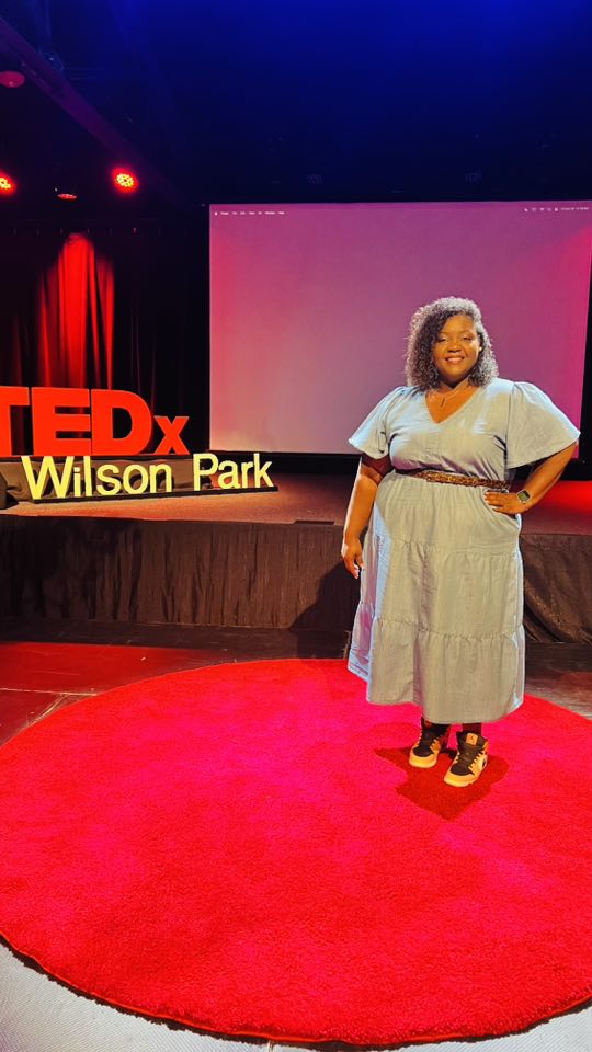 Woman standing on red carpet in front of TEDx Wilson Park sign and screen, smiling, wearing a light-colored dress and sandals.