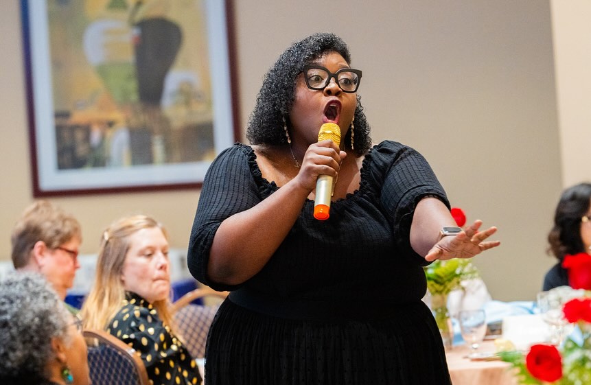 Woman speaking into a microphone at a gathering, with seated audience members in the background.