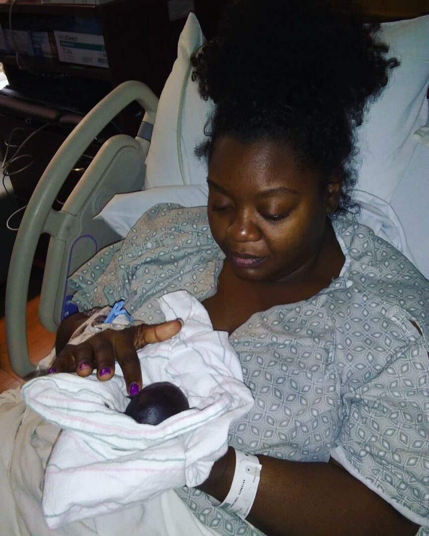 Woman in hospital bed holding newborn baby wrapped in white blanket, hospital room background.