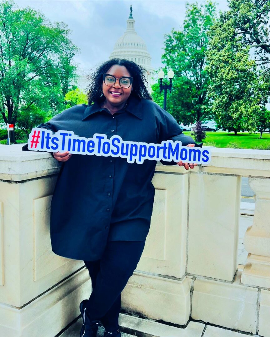 Smiling woman holding a sign with hashtag #ItsTimeToSupportMoms in front of a white building and green trees.