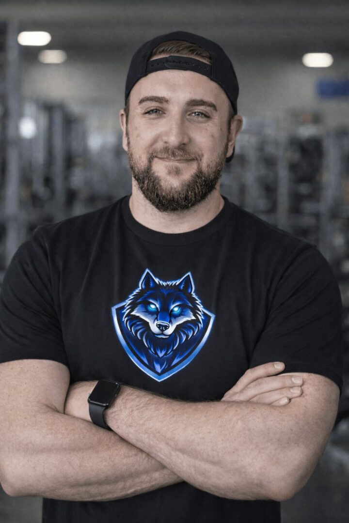 Man with beard wearing a black cap and T-shirt with a wolf logo, arms crossed, in a gym setting.
