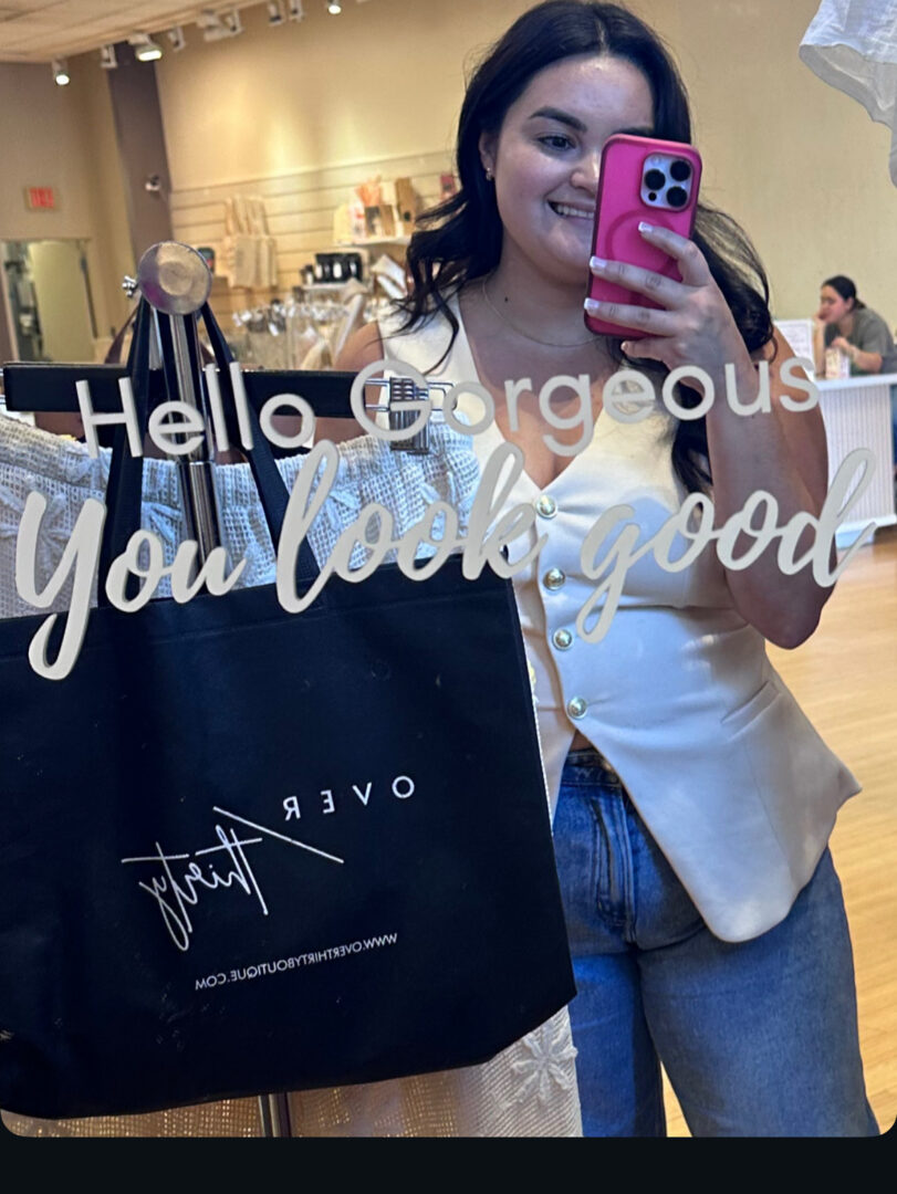 Woman taking mirror selfie with pink phone, smiling, in a store or cafe, holding a large black shopping bag.