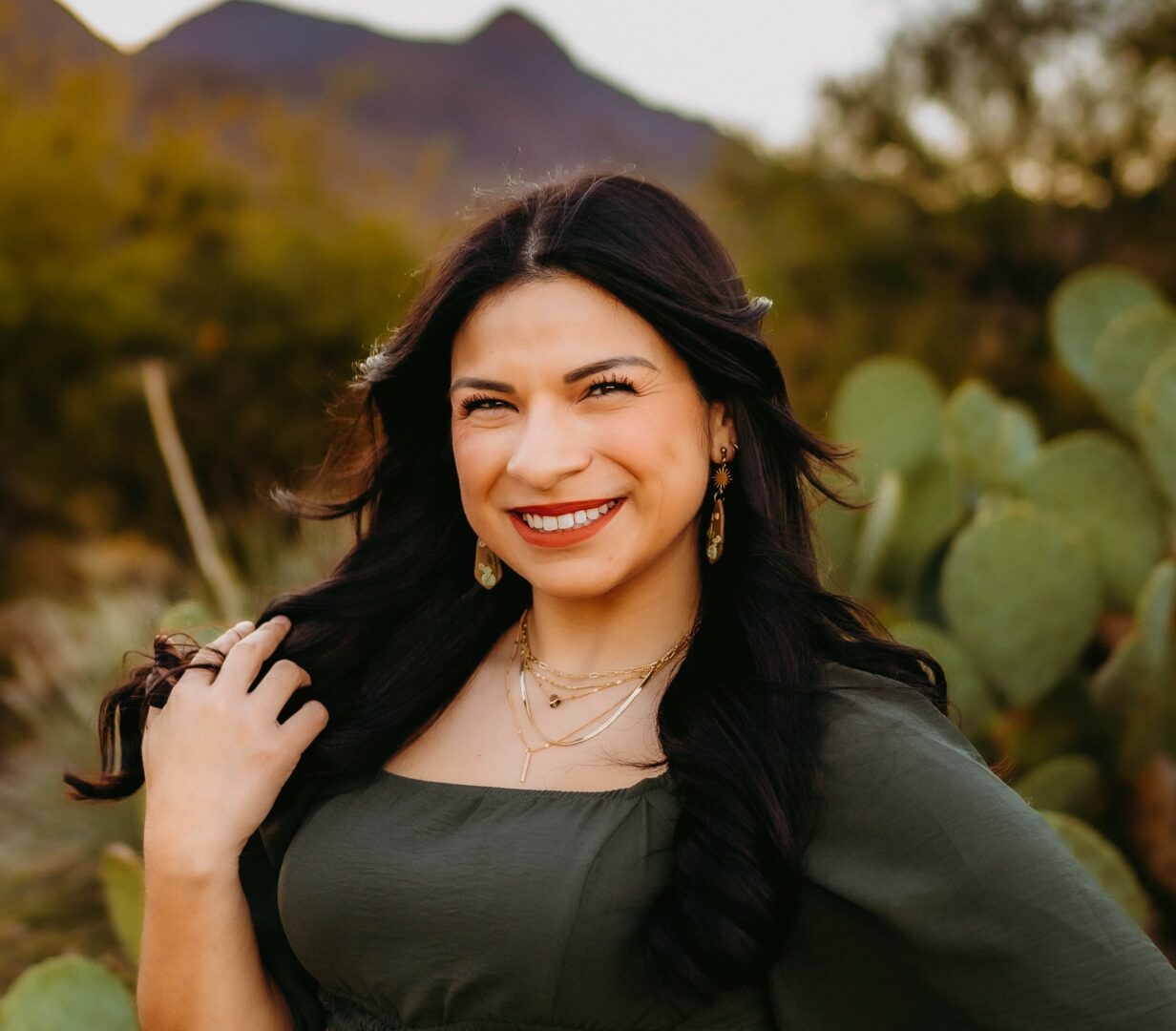 Smiling woman with long dark hair outdoors, mountains and cactus in background.