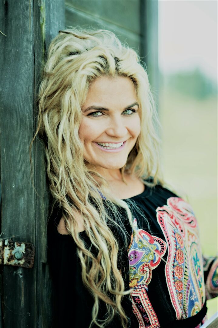 Woman with long, curly blonde hair smiling outdoors, leaning against a wooden structure, wearing a colorful top.