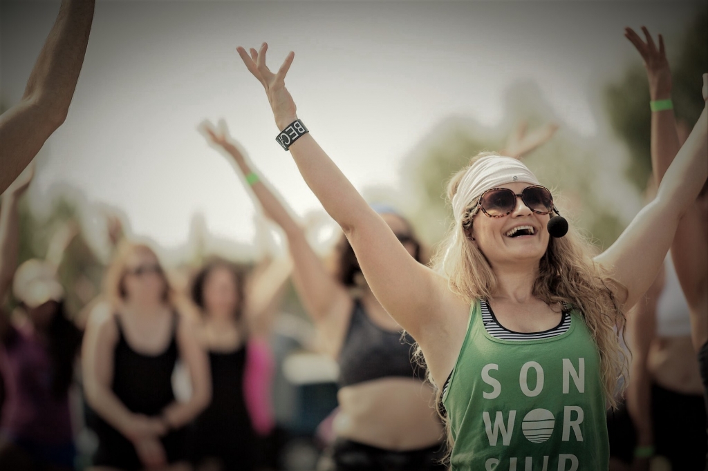 Woman with sunglasses and headscarf raising arms at outdoor event, smiling, with group of people in background.