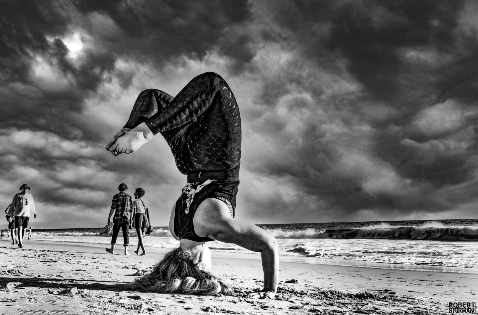 Person performing a headstand on the beach with cloudy sky and ocean in background.