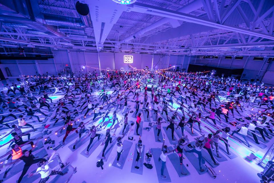 Large indoor event with many people practicing yoga on mats under purple lighting.