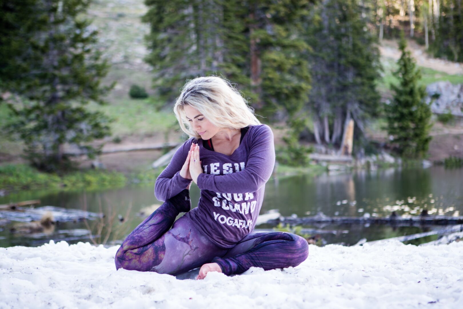 Woman sitting cross-legged on sand near a lake with trees in the background, practicing yoga or meditation outdoors.