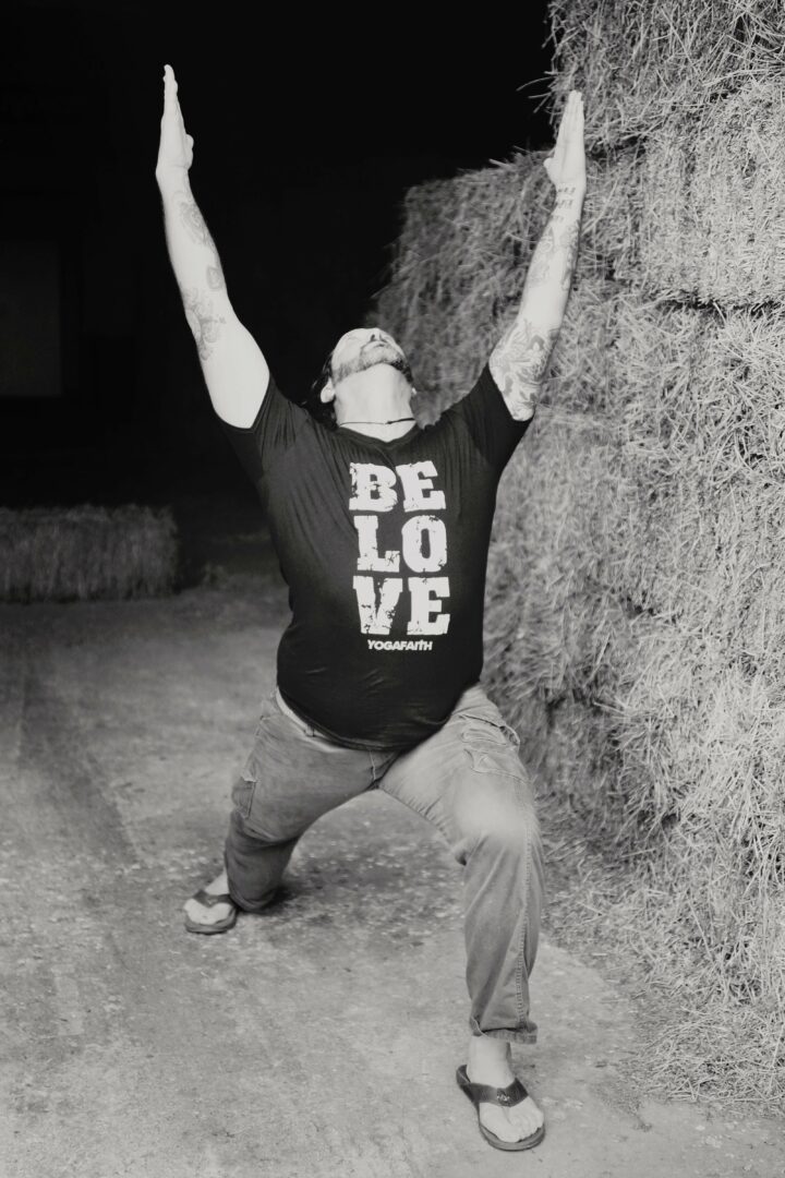 Person standing with arms raised, leaning against a textured wall, wearing a black T-shirt and jeans.