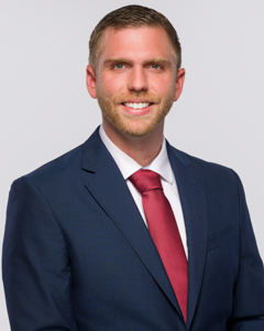 Man in a navy suit with a red tie, smiling, against a light gray background.