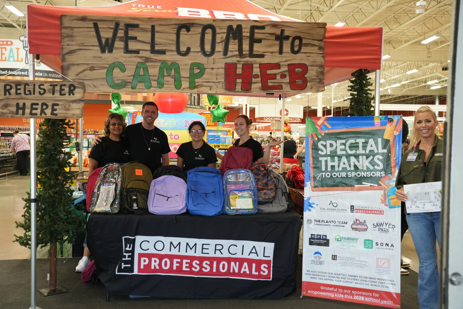 Group of five people standing behind a table with backpacks at a camp event, with a welcome banner and sponsor sign nearby.