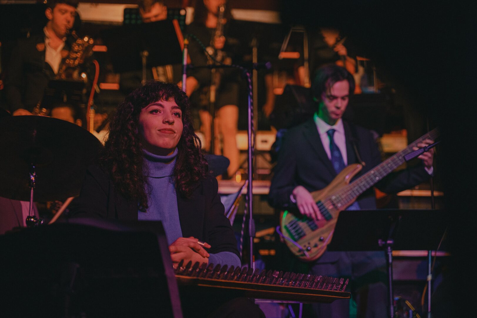 Two musicians perform on stage, one playing keyboard and the other playing bass guitar, with an orchestra in the background.