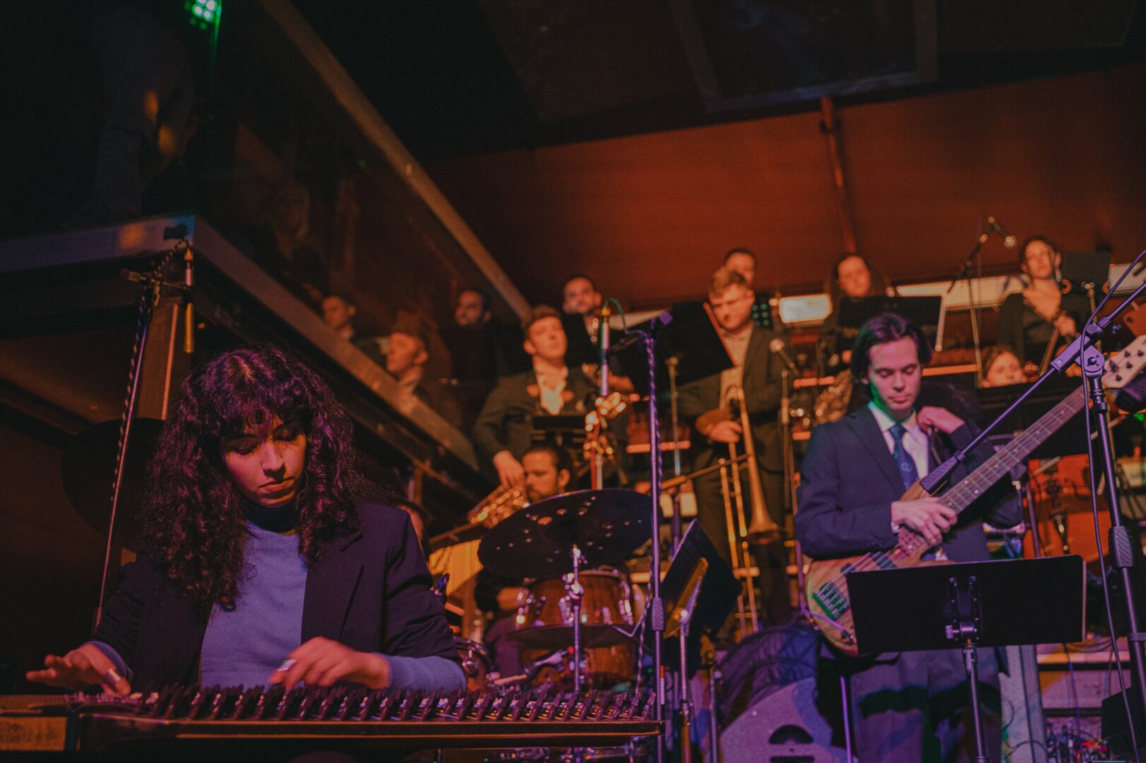 Orchestra performing on stage with musicians playing instruments, including a keyboard and guitar, in a dimly lit venue.